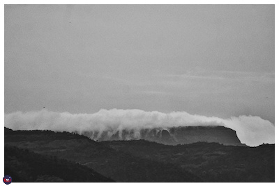 A black and white photograph of a hill covered with clouds, where the clouds are immitating giant waves.